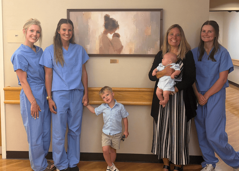 Three nurses in blue surgical scrubs, a woman in a black sweater and striped pants holding a baby, a young boy in a blue shirt and khakis stand posed in front of a illustration of a woman and a baby
