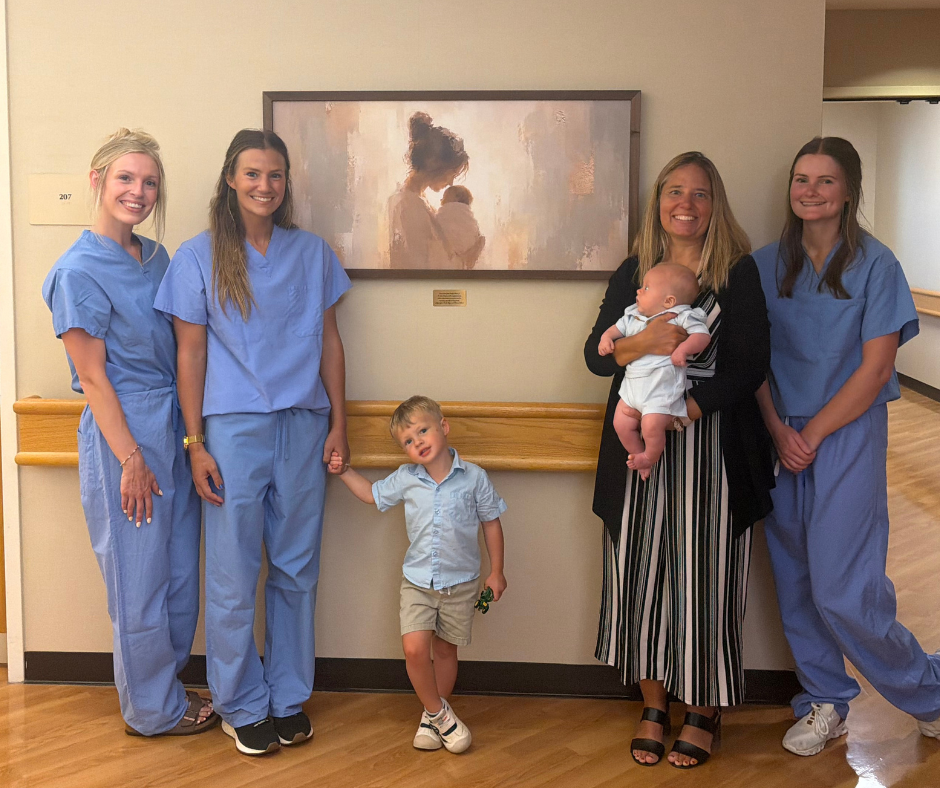 Three nurses in blue surgical scrubs, a woman in a black sweater and striped pants holding a baby, a young boy in a blue shirt and khakis stand posed in front of a illustration of a woman and a baby