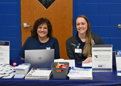 Two women sit at a table behind laptops and paper materials