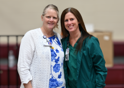 A woman in a white sweater and white and blue dress and a woman in green scrubs stand posed together.
