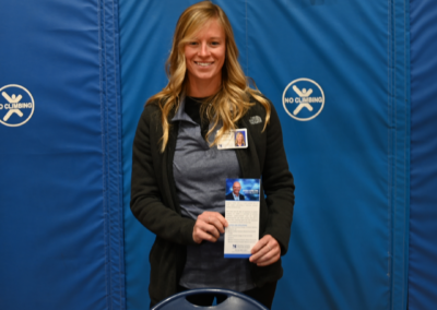 A woman in a polo and sweater stands in front of a blue background holding a piece of paper