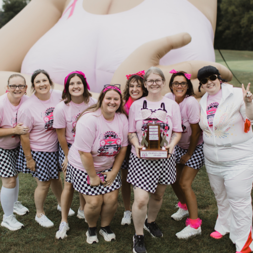 Women in pink t-shirts and black and white checkered skirts stand posed for a photo holding a trophy