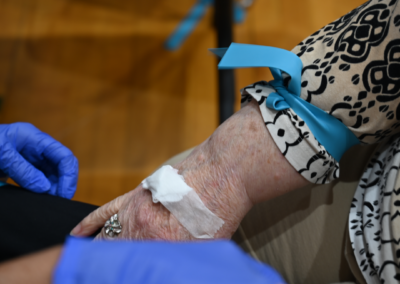 A pair of blue gloves is worn by a woman assisting a woman with a bold print top in a blood draw