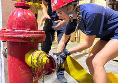 Girl in a navy outfit and red helmet with braid attaches a yellow hose to a red firehydrant