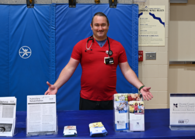 A man in a red scrub top and black pants stands posed behind a table