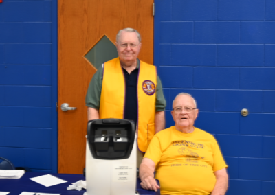 Two older men sit and stand next to one another, posed with a device to examine vision.