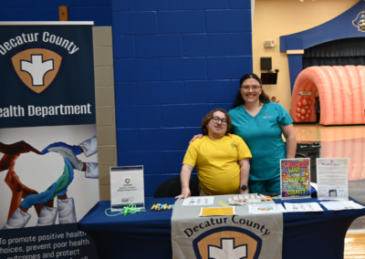 A man in a yellow shirt and woman in a blue scrub top stand behind a table that says "Decatur County Health Department"