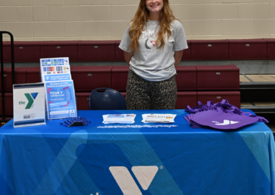 A woman with long red hair and a gray shirt stands behind a blue tablecloth that says "the Y"