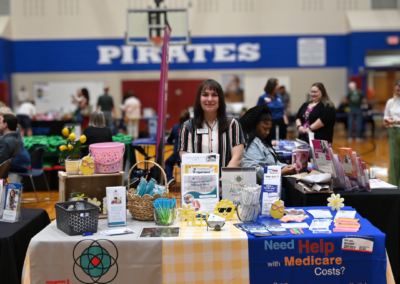 A woman stands behind a yellow gingham table with two signs, one is for "Thrive Alliance" the other for "Indiana SHIP"