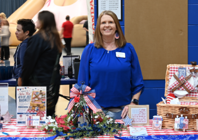 A woman with red hair and a royal blue tshirt stands behind a red gingham table with various red white a blue items on it