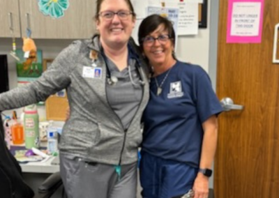 Two Women with brown hair stand posed together in front of a desk