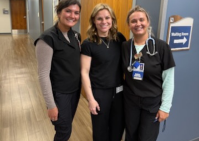 Three young women in black scrubs stand posed in front of a blue wall