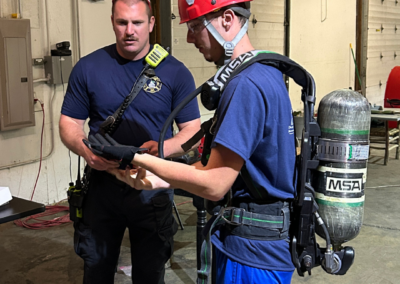 Young man with red helmet and blue outfit wears and oxygen tank next to a firefighter in a navy top and black pants