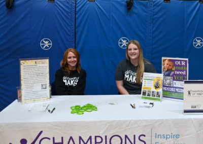 Two women in black shirts sit behind a white table with paper materials and signs, reading "Champions of Youth"