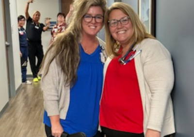 Two blond women in light sweaters with bold tops stand posed in front of a hallway