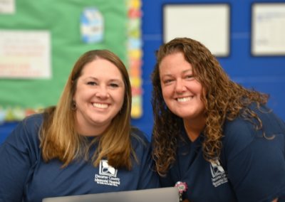 two women with brown hair and navy t-shirts stand posed next to one another.