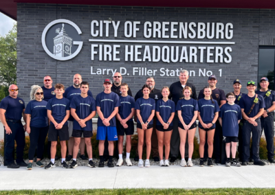 Children in navy t-shirts stand posed with police, fire, and emergency service workers in front of a fire station
