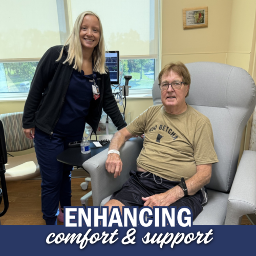 A woman with blonde hair and navy blue scrubs stands posed with a man seated in a gray recliner with red hair, a tan shirt, and black shorts.